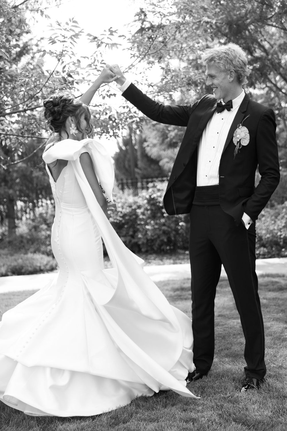 A bride in a flowing white gown twirls while dancing with her groom in a black tuxedo outdoors in black and white.