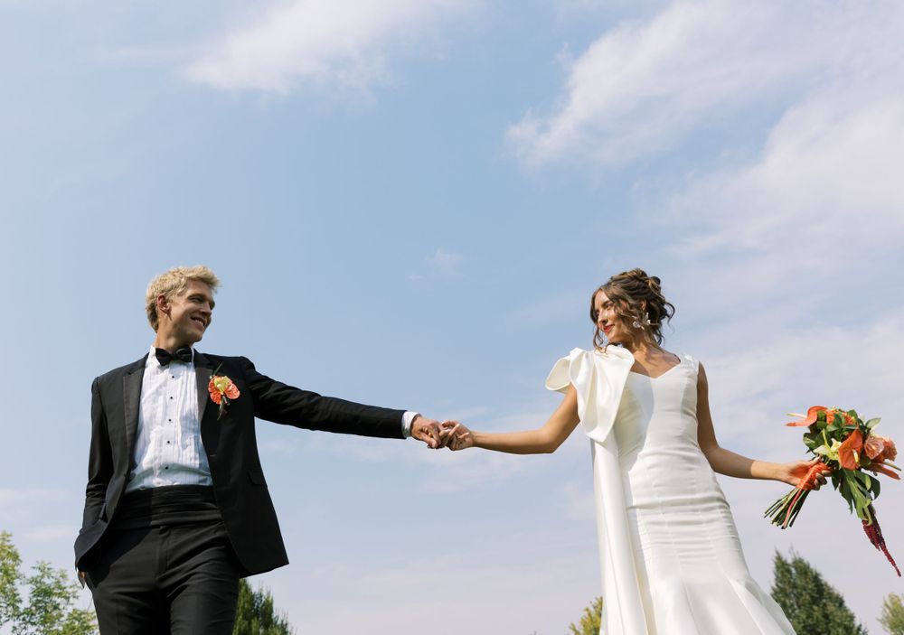 Happy newlyweds hold hands against a blue sky on their outdoor wedding day, with bride holding orange bouquet.