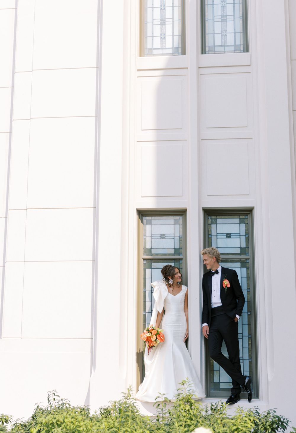 A couple stands outside a white church building with tall windows, the bride holding an orange bouquet on their wedding day.