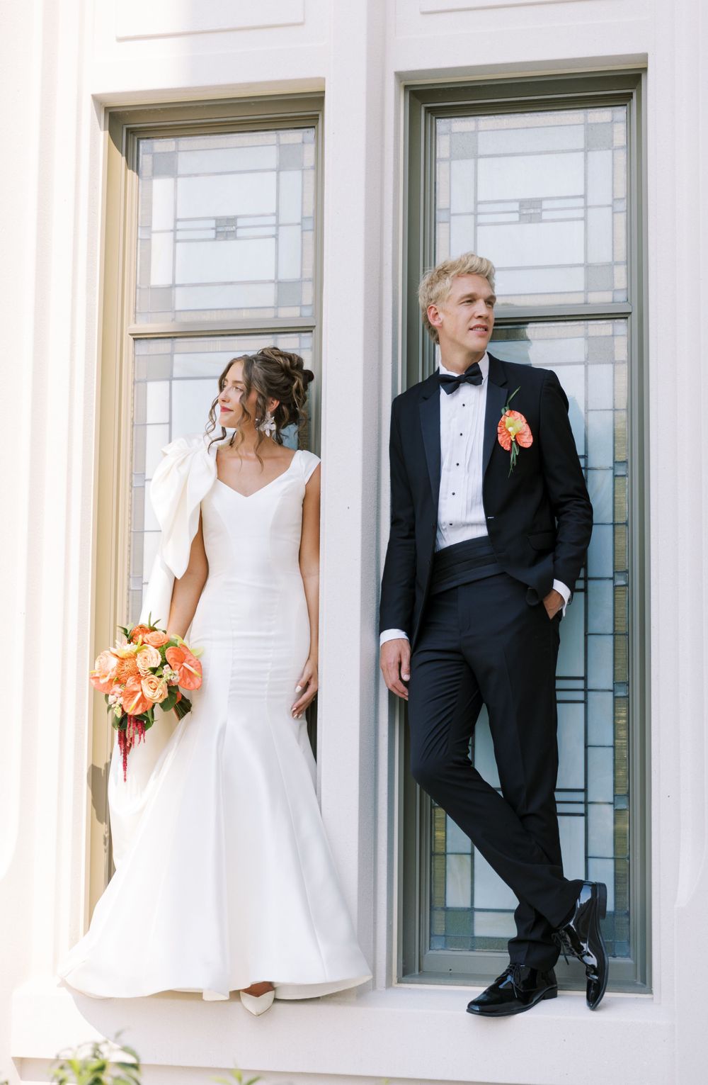 Elegant wedding couple poses by window frames, bride in fitted white gown holds peach bouquet while groom wears classic tuxedo.