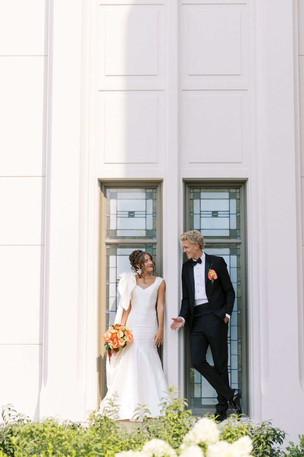 Elegant couple in wedding attire share a tender moment against a white church building with tall windows.