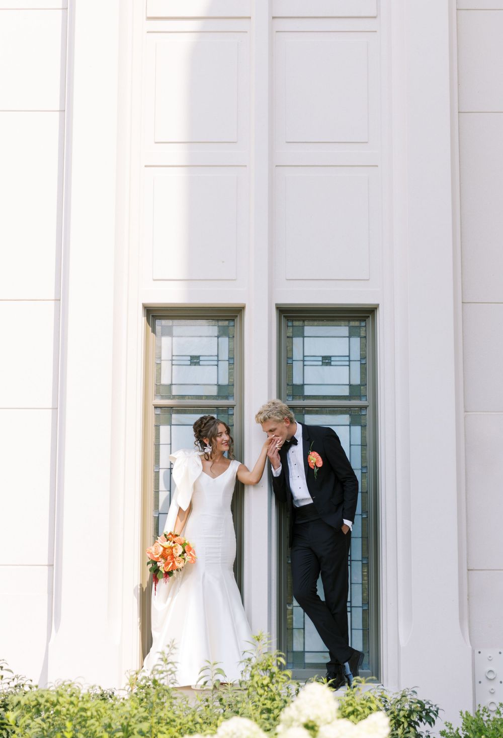 A couple poses together casually against white pillars in formal wedding attire, with the bride holding bright orange flowers.