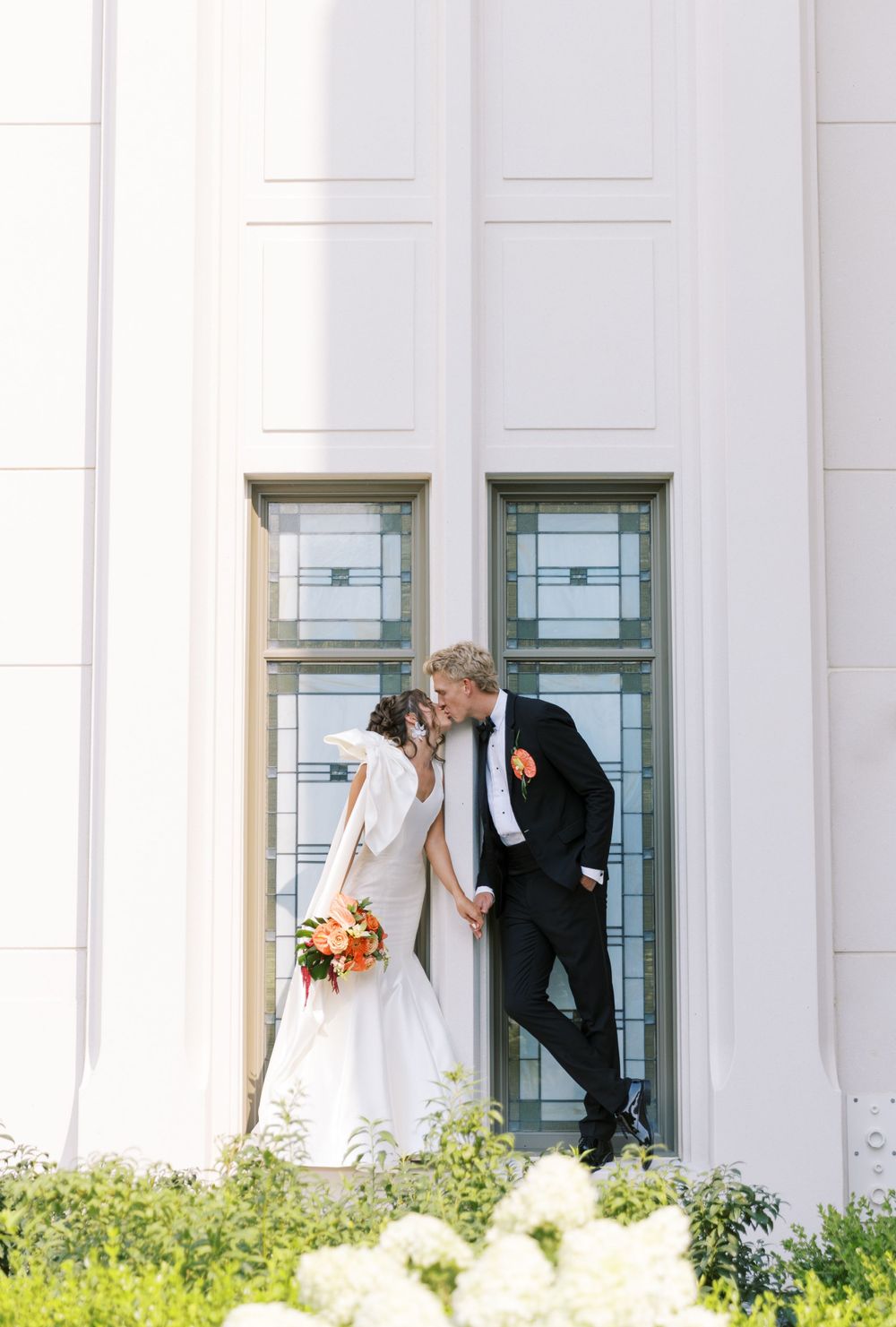 A couple in wedding attire shares a tender moment against a white building with decorative windows and greenery.