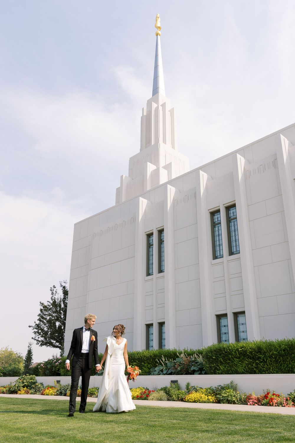 A couple stands on manicured lawn in front of a white LDS temple with a tall spire against a cloudy sky.
