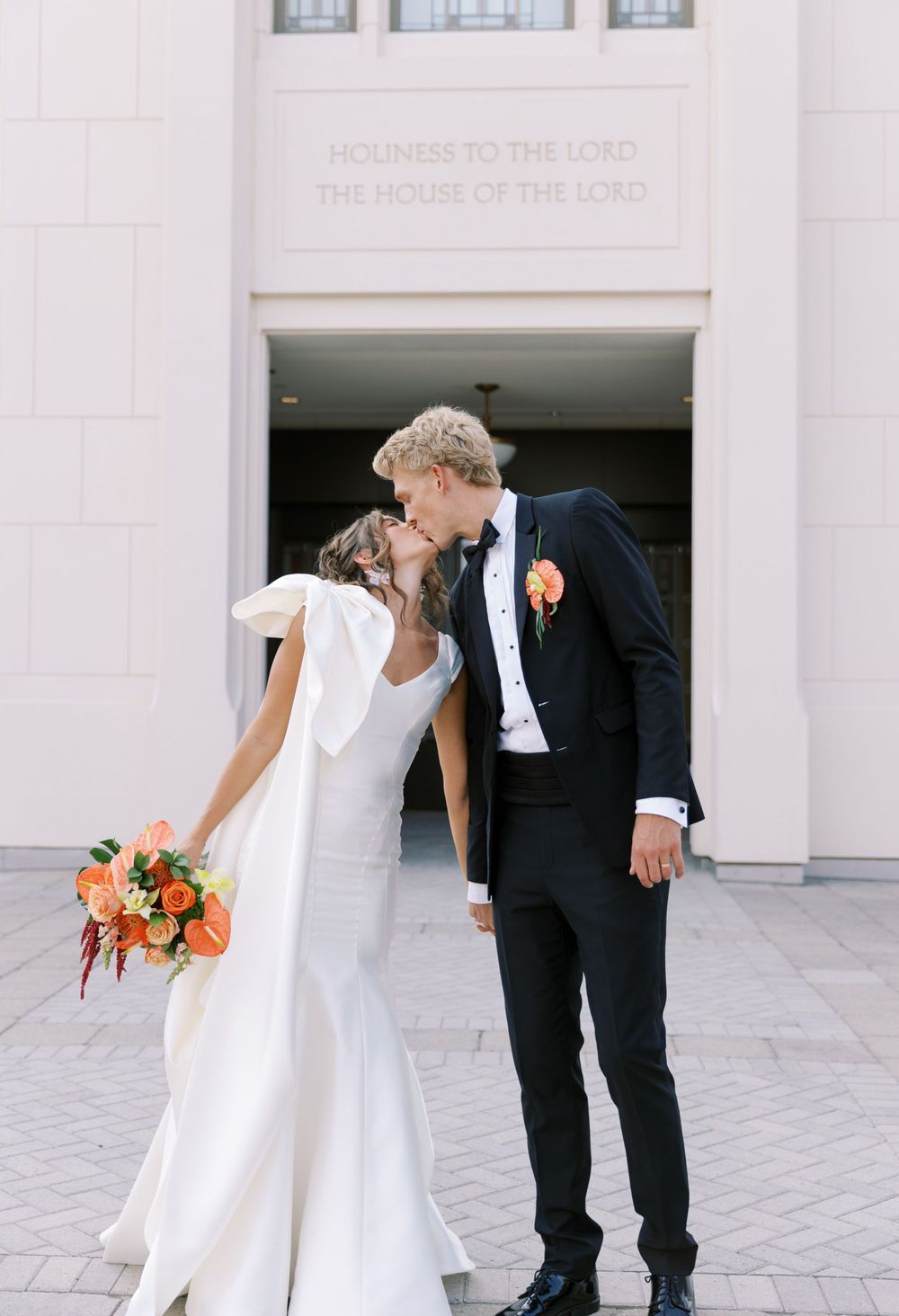A bride in a white dress and groom in a black suit share a tender moment outside a church entrance.
