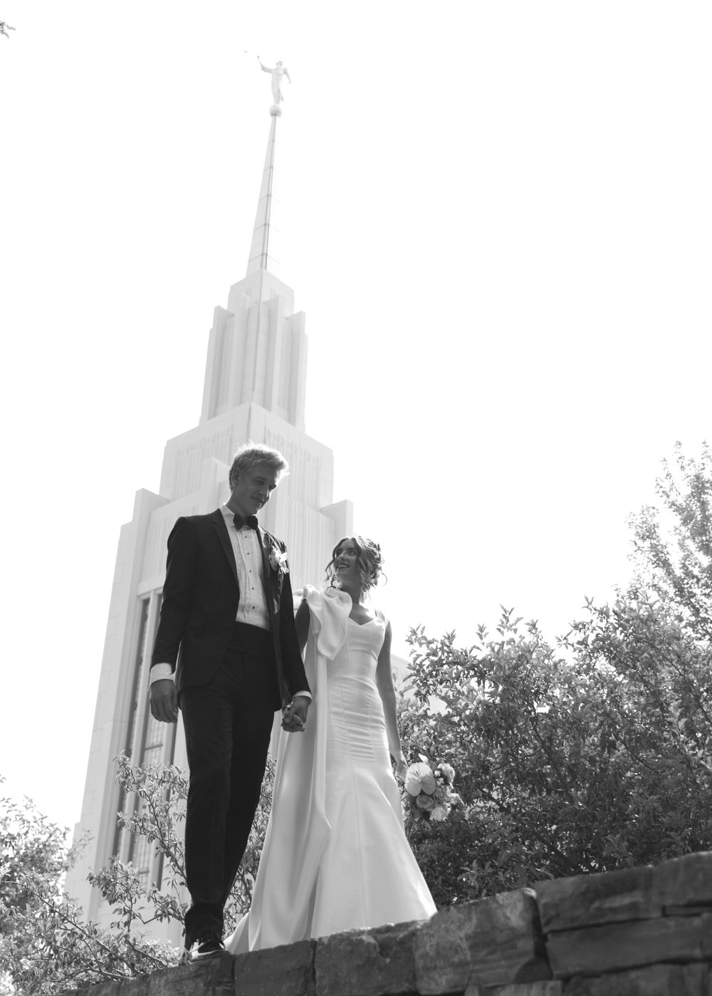 Black and white wedding photo of a couple standing in front of a tall LDS temple spire on their special day.