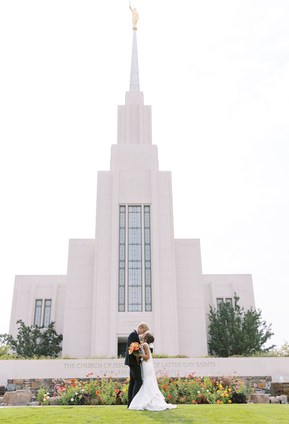 A wedding couple poses in front of a white LDS temple with blooming flowers in the foreground.