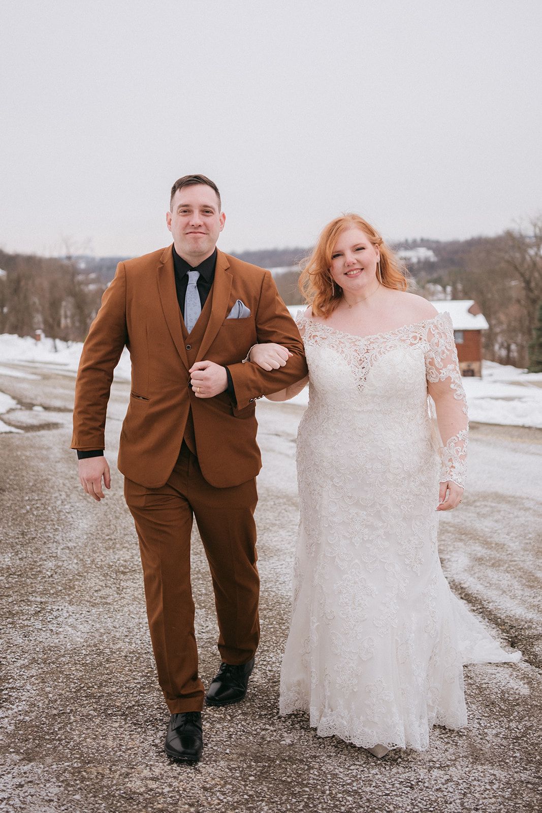 Two people in wedding attire share a joyful moment together outdoors while looking down and smiling warmly.
