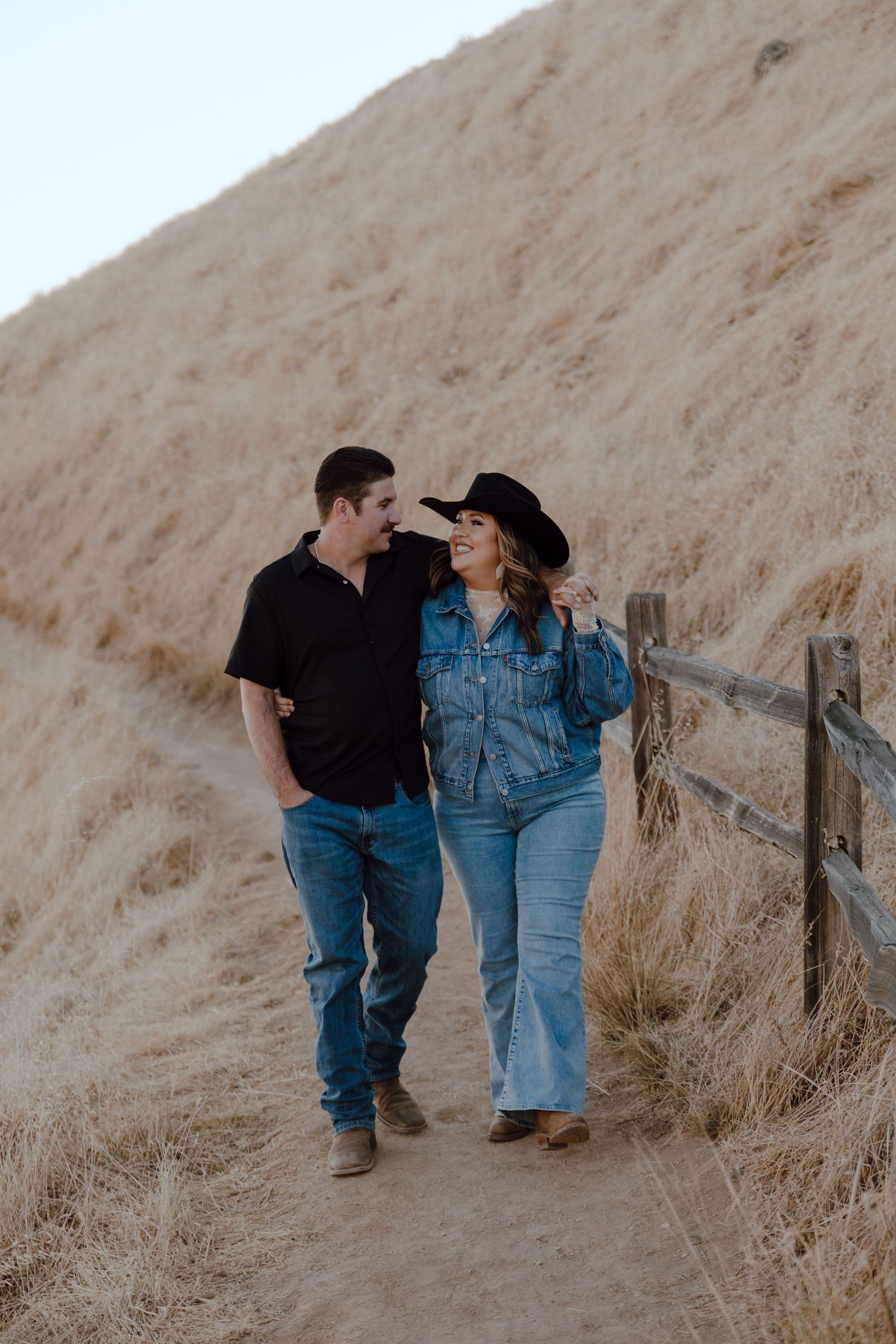 A couple in denim walking together on a dirt path along a golden hillside with a wooden fence at sunset.