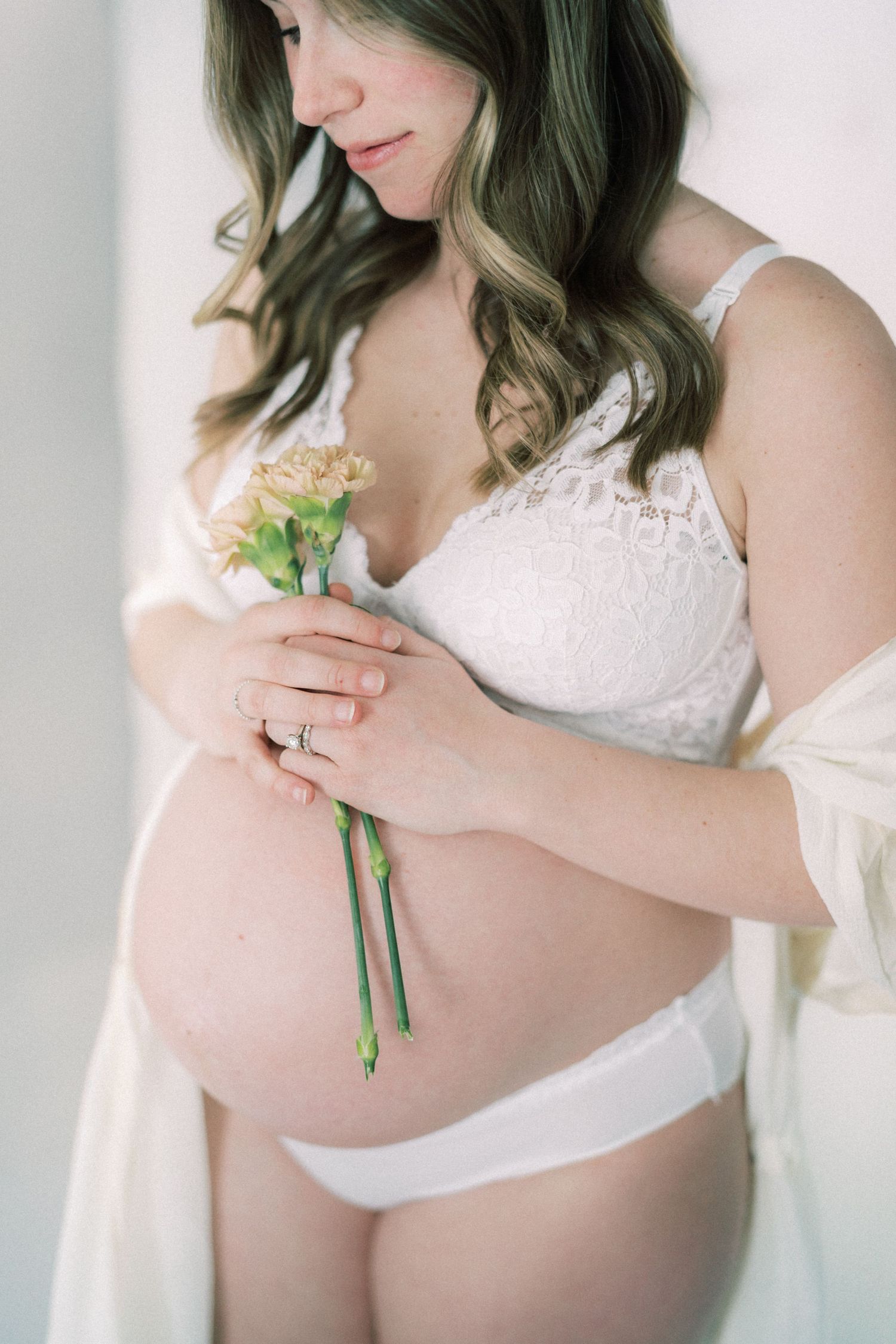 Ethereal maternity portrait showing woman in white lace holding delicate flowers against her belly.