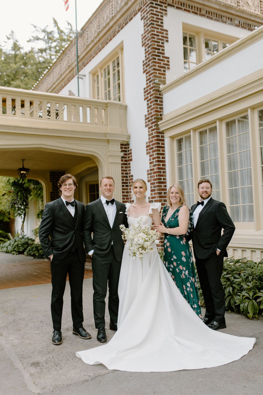 Wedding party in formal attire poses outside elegant brick and white mansion with grand pillars and windows.