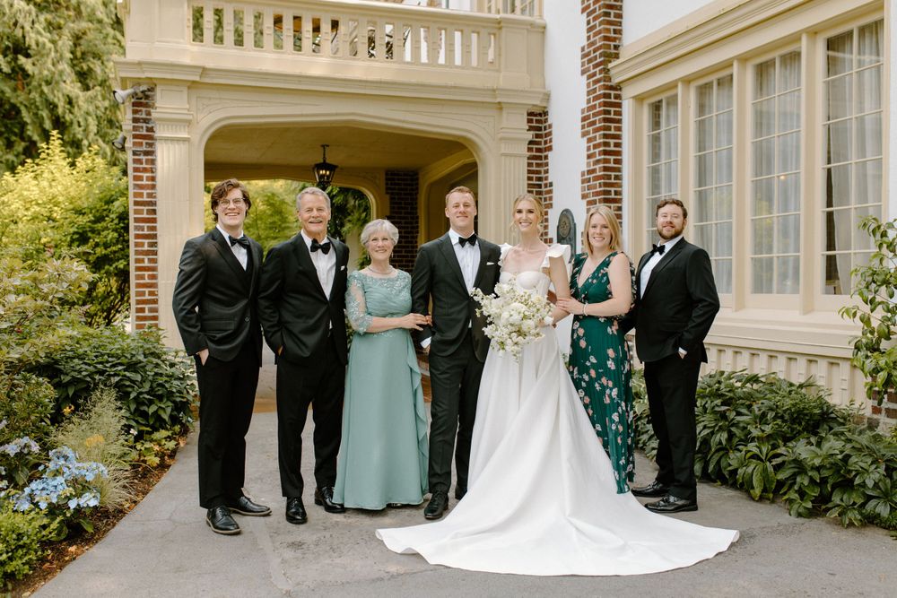 A formal wedding party group stands together outside an elegant stone mansion with manicured gardens.