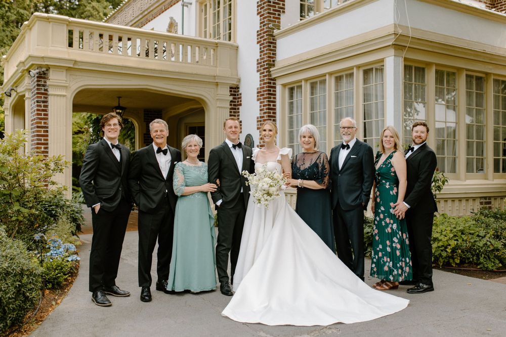 A wedding party in formal attire poses outside a grand white mansion with columns and palm trees.