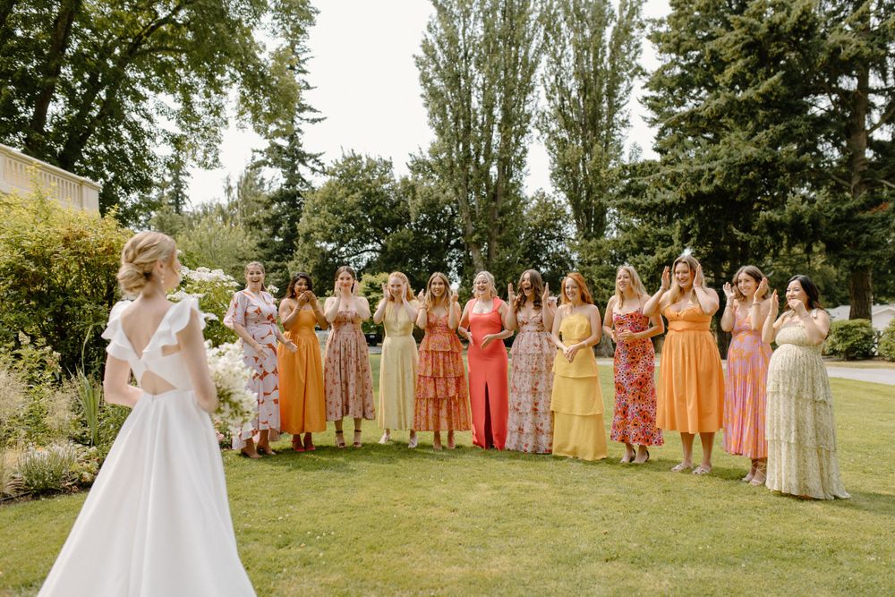 A wedding party stands in a garden setting wearing an array of warm-colored dresses in yellow, orange and peach tones.