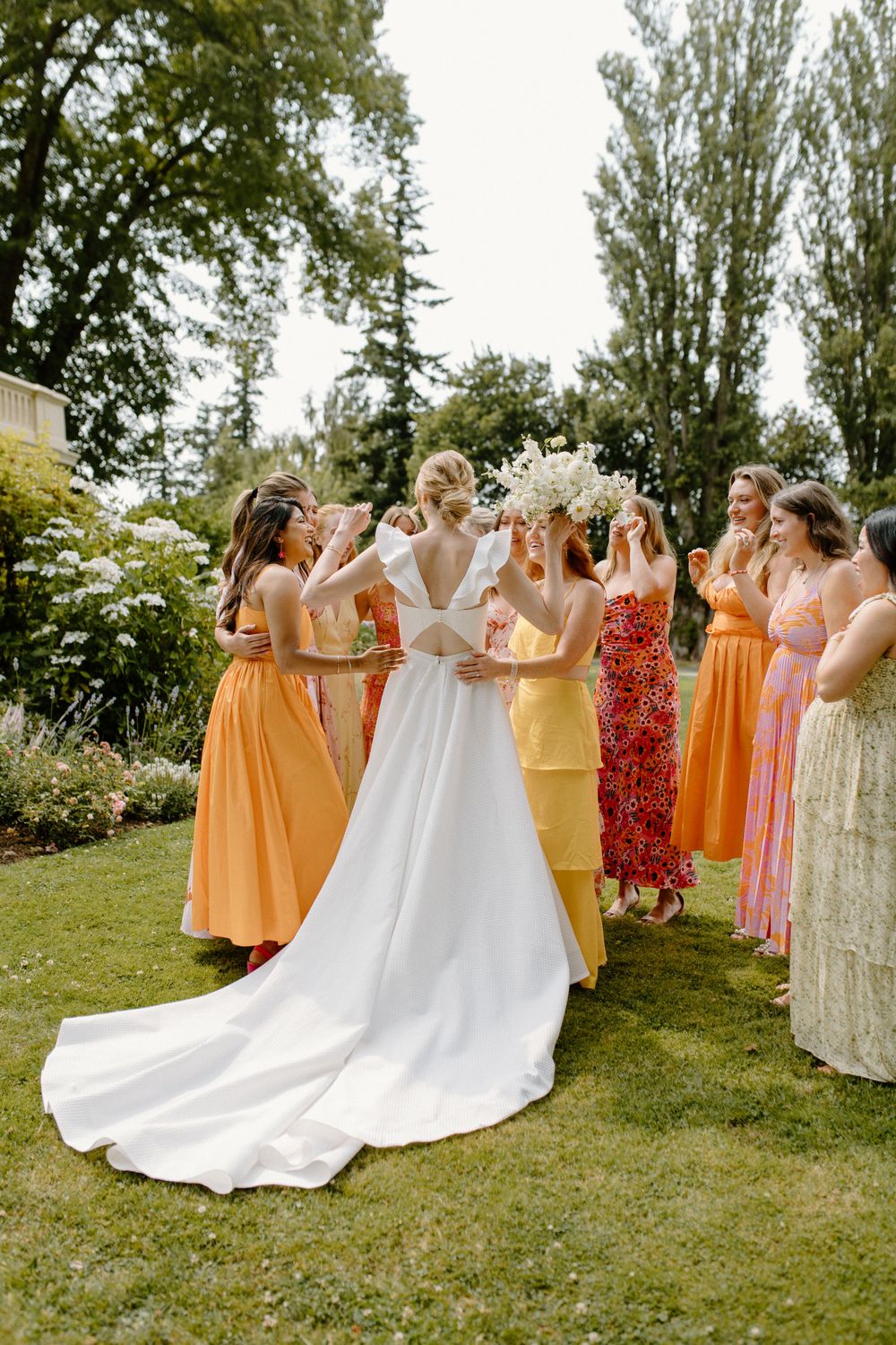 A wedding party in colorful dresses gathers on a lawn surrounded by trees during an outdoor summer celebration.