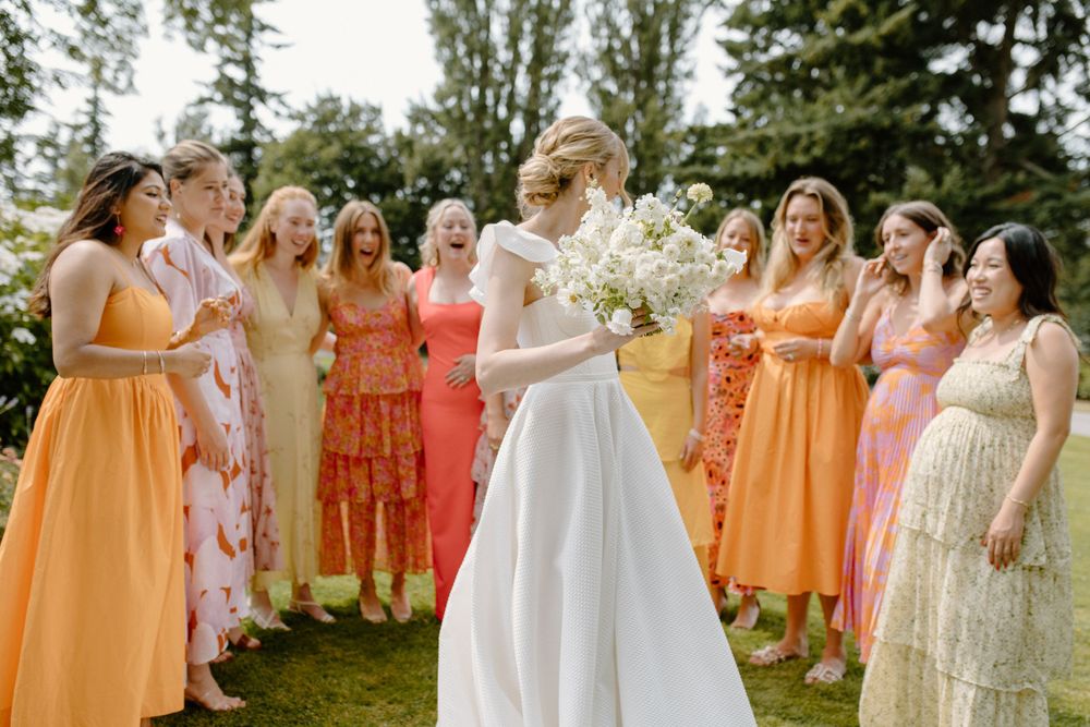 Wedding party gathers outdoors in coordinating peach, coral and orange dresses on a sunny summer day.