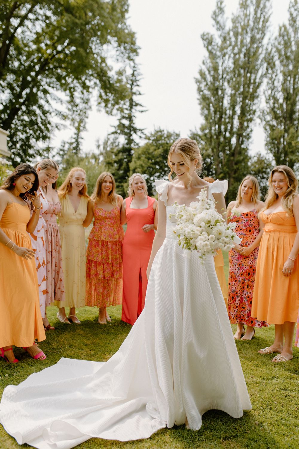 Bride in elegant white gown stands with bridesmaids wearing colorful orange and floral dresses on sunny outdoor lawn.