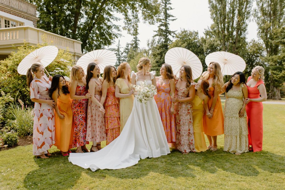 Bridesmaids holding white parasols pose with bride in flowing white gown on sunny lawn during summer wedding celebration.
