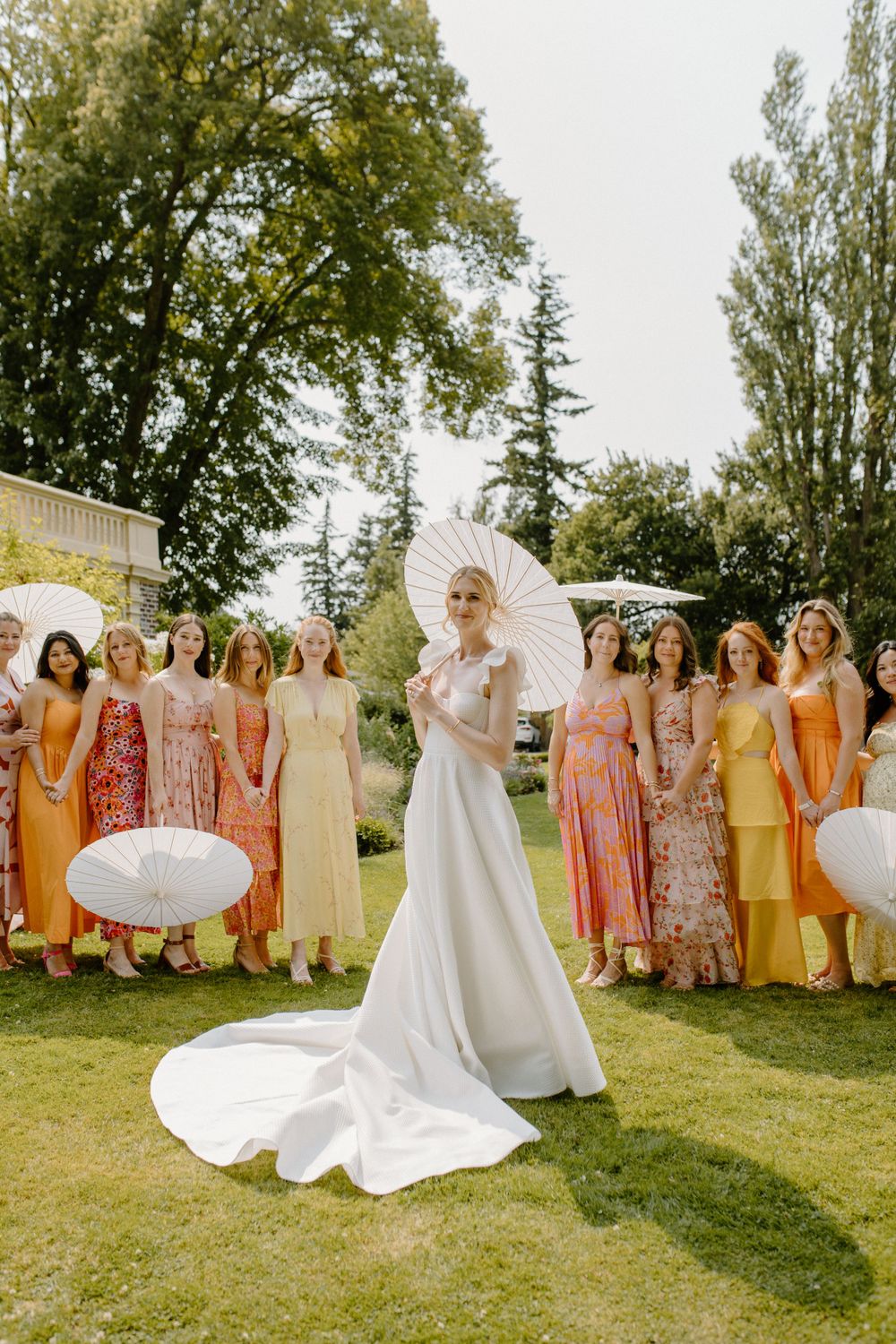 A wedding party poses on a sunny lawn with bridesmaids in warm pastel dresses and the bride in an elegant white gown with parasols.