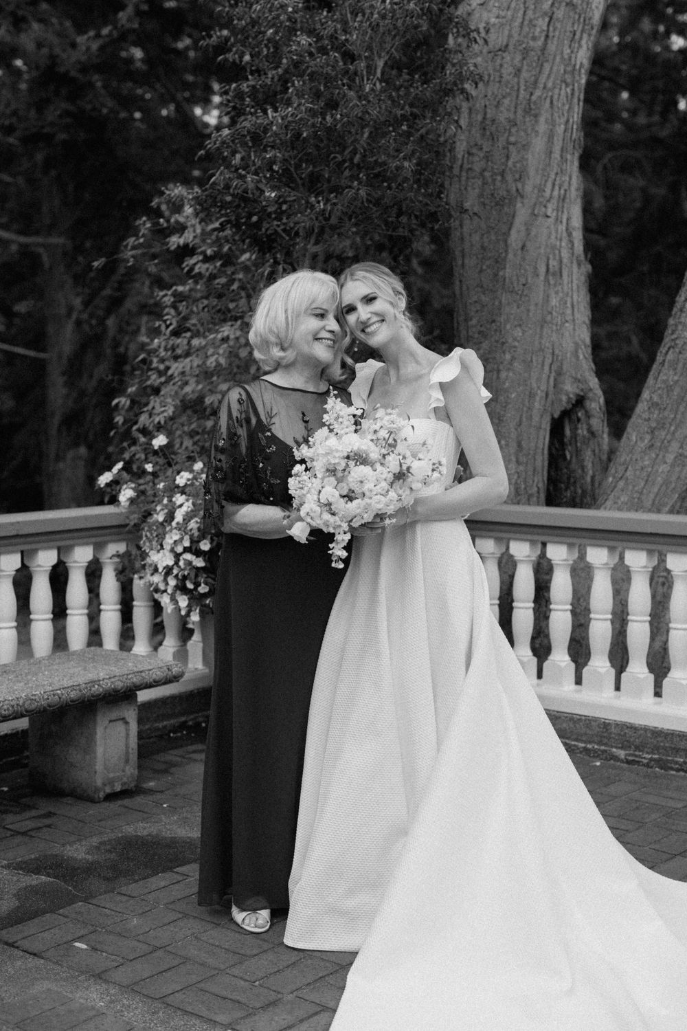 Black and white wedding photo of two people sharing a tender moment on a balcony with stone balustrades and trees in background.