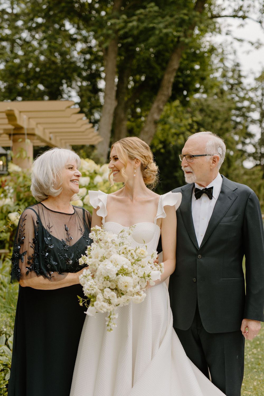 Wedding moment captured outdoors with white floral bouquet and formal attire against garden pergola background.
