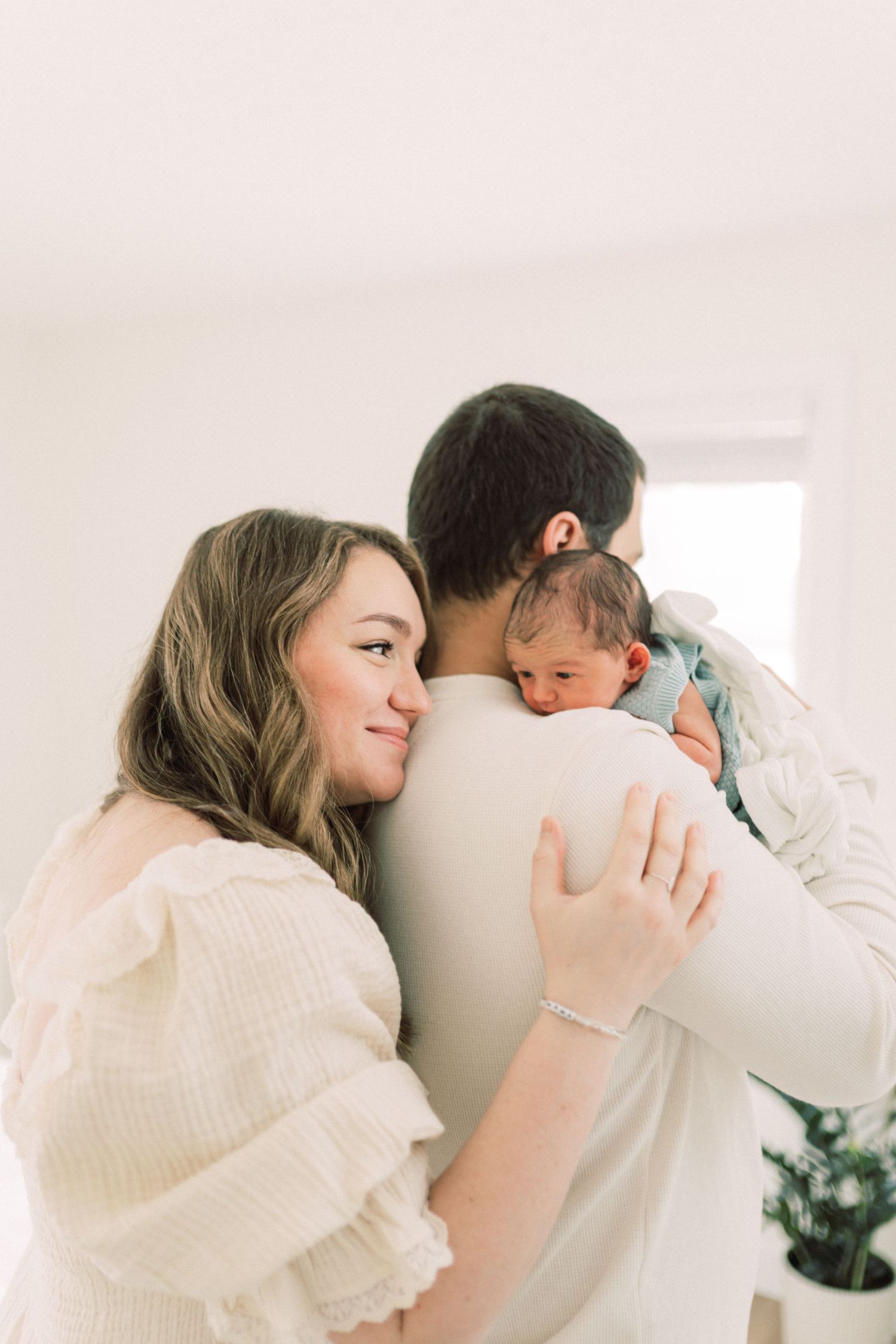 Tender family moment captured in soft natural light with parents embracing newborn baby in white room.