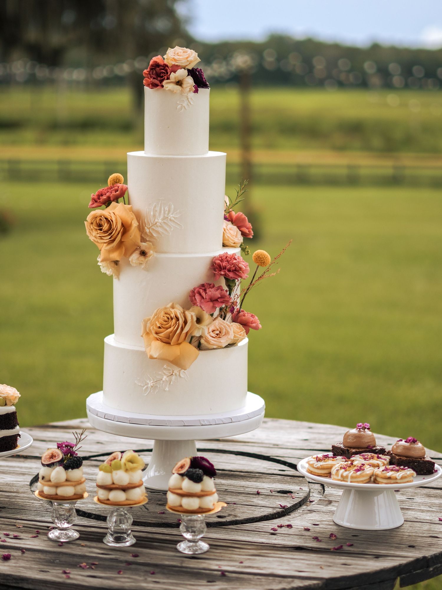 Elegant four-tier white wedding cake decorated with peach and pink roses on display with desserts at outdoor reception.
