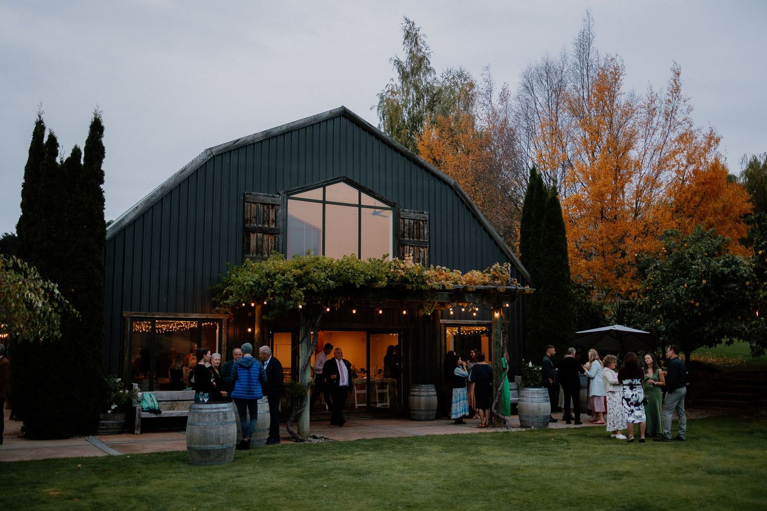 A rustic navy blue barn venue decorated with string lights hosts an evening gathering surrounded by autumn trees.