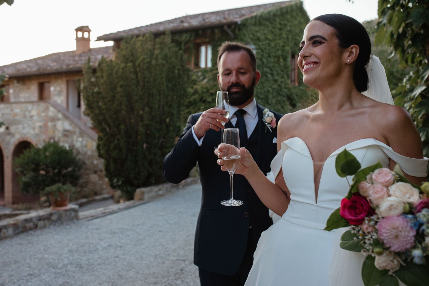 Newlyweds share a laugh while holding champagne glasses outside an elegant stone villa in Italy during their wedding celebration.