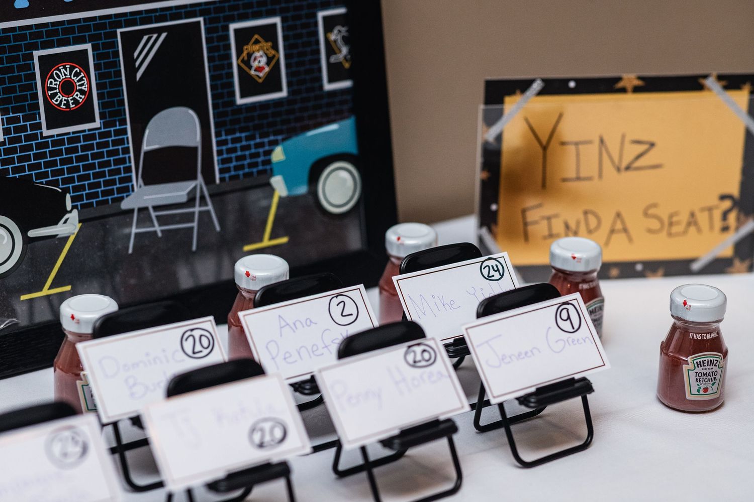Small black folding chairs with white name cards arranged in a row next to a jar on a table against a decorated wall.