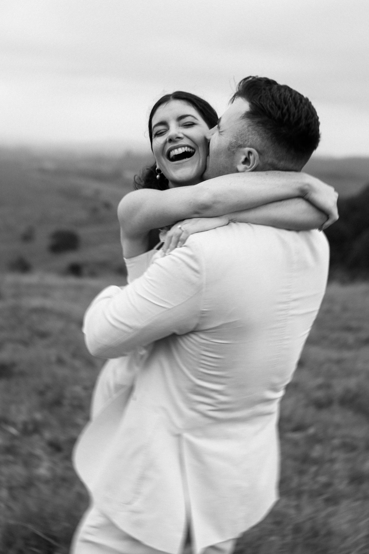 An intimate black and white portrait of a couple embracing in a pastoral landscape.