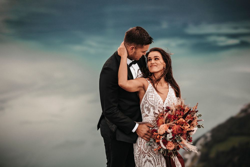 Dramatic wedding portrait against moody sky with bride holding vibrant autumn bouquet on mountainous clifftop.