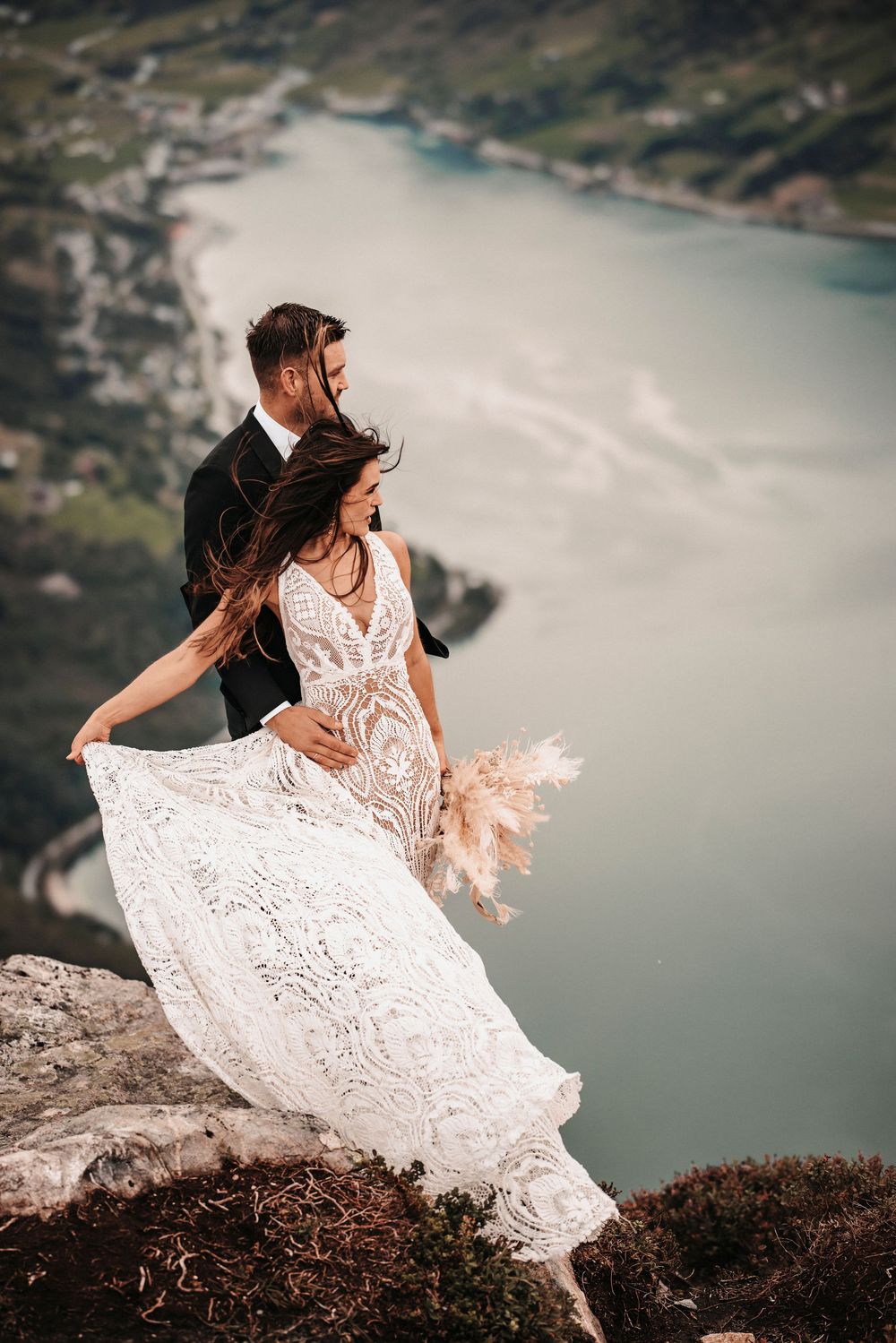 A romantic wedding photo featuring a lace dress flowing in the wind on a cliff overlooking a scenic lake.