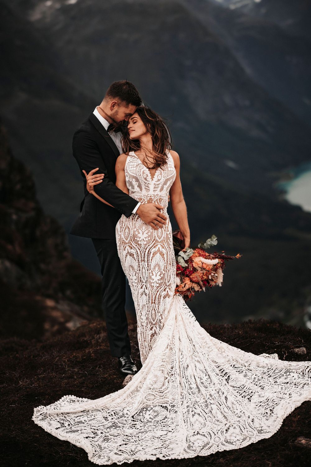 A wedding couple embraces on a mountain overlook with a dramatic lace train wedding dress and autumn bouquet.