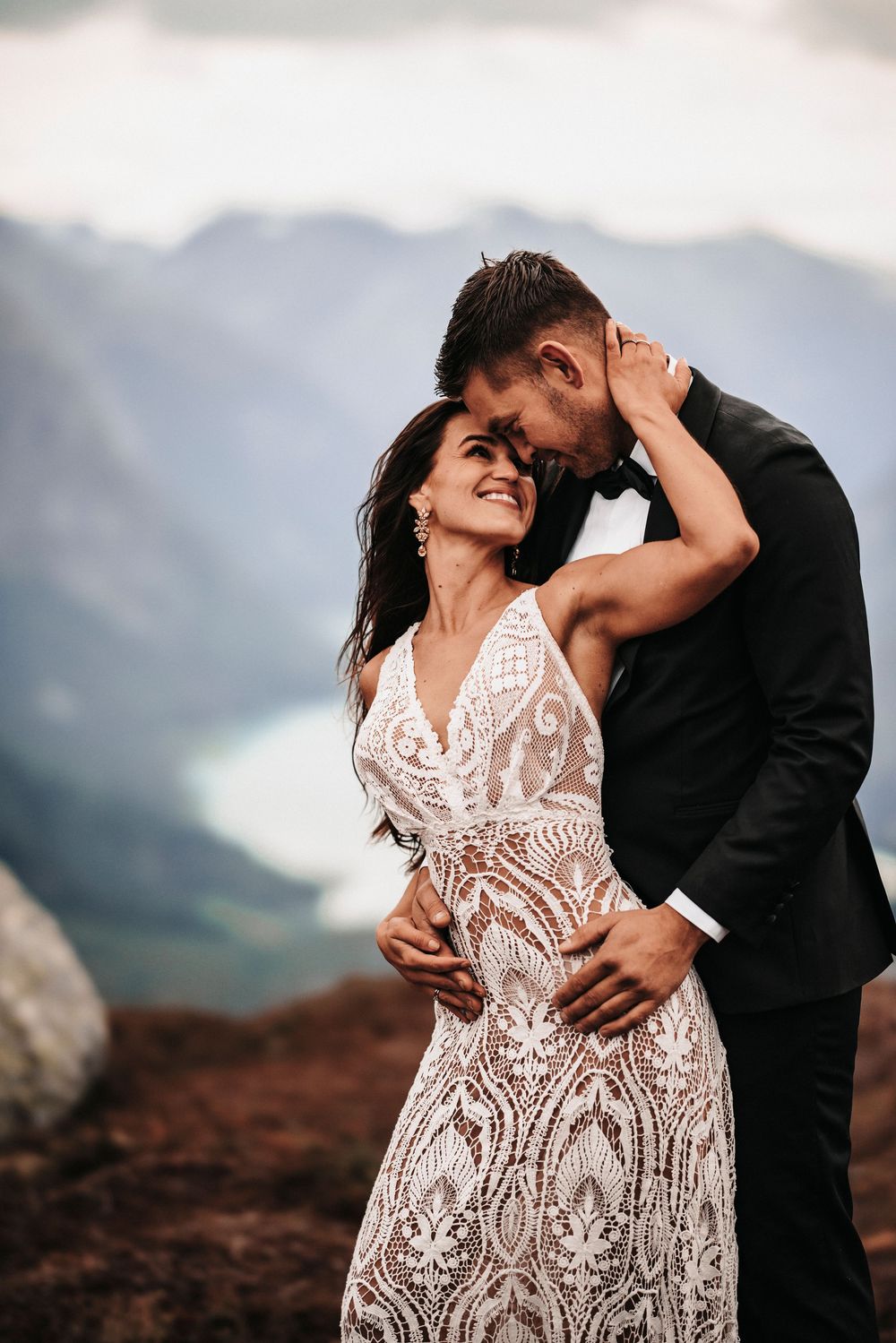 A couple in wedding attire embrace on a mountaintop with a dramatic mountain landscape and lake in the background.