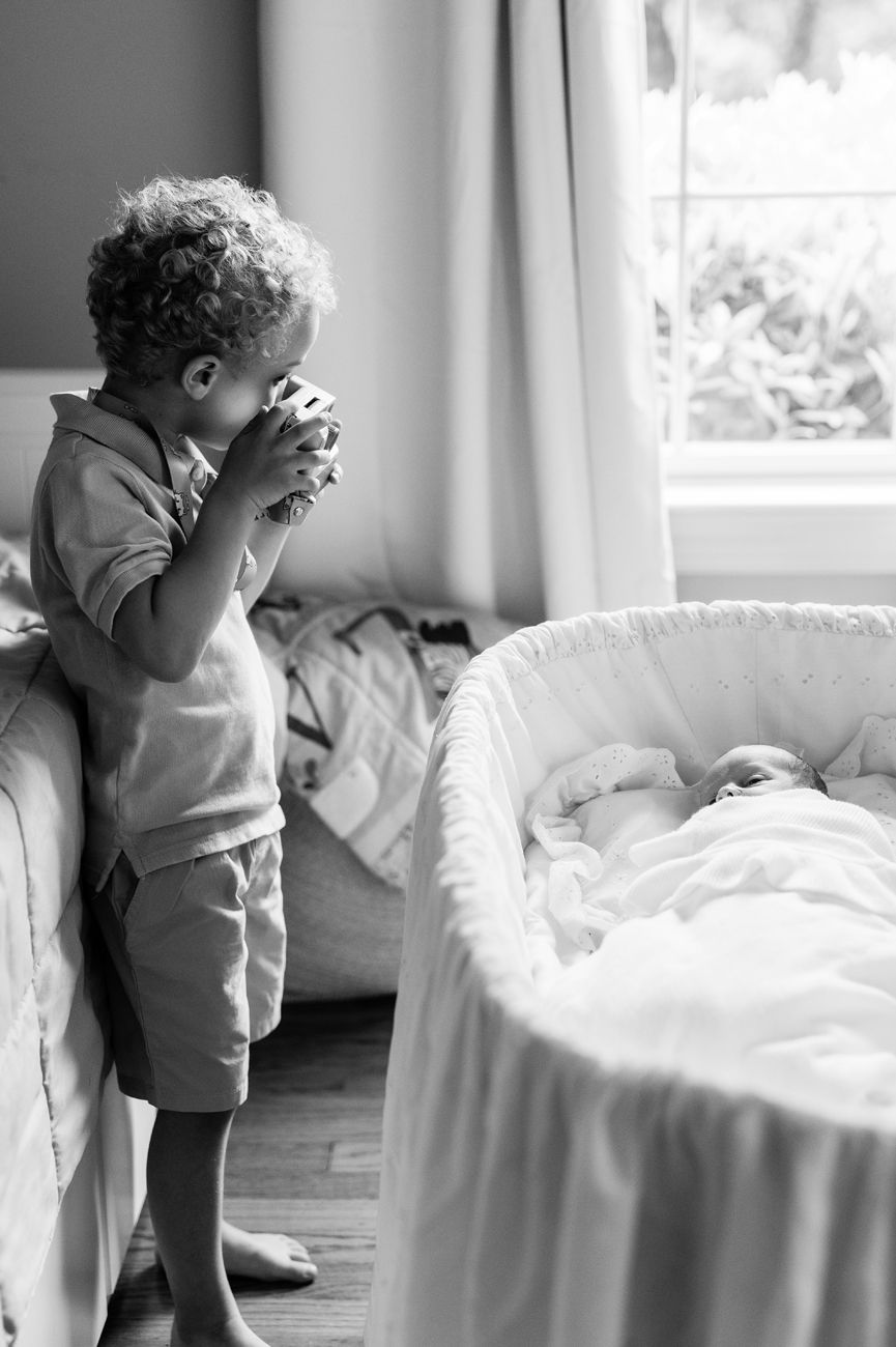 A toddler peeks over the edge of a white bassinet in a softly lit bedroom.