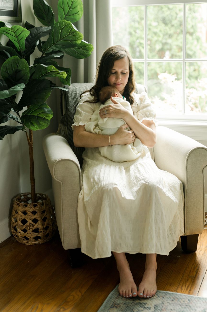 A person in a white dress sits in a cream armchair by a window with a fiddle leaf fig plant and woven basket nearby.
