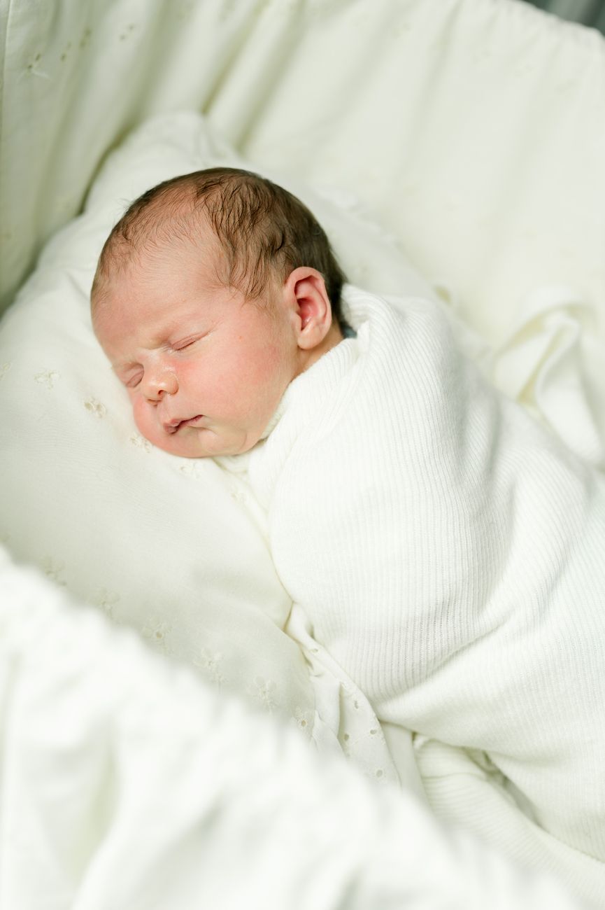 A sleeping newborn swaddled in white blankets rests peacefully on white bedding.