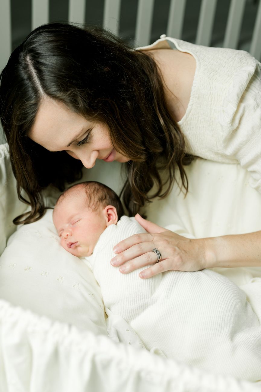 A parent in a cream sweater tenderly watches over their sleeping newborn swaddled in white bedding.