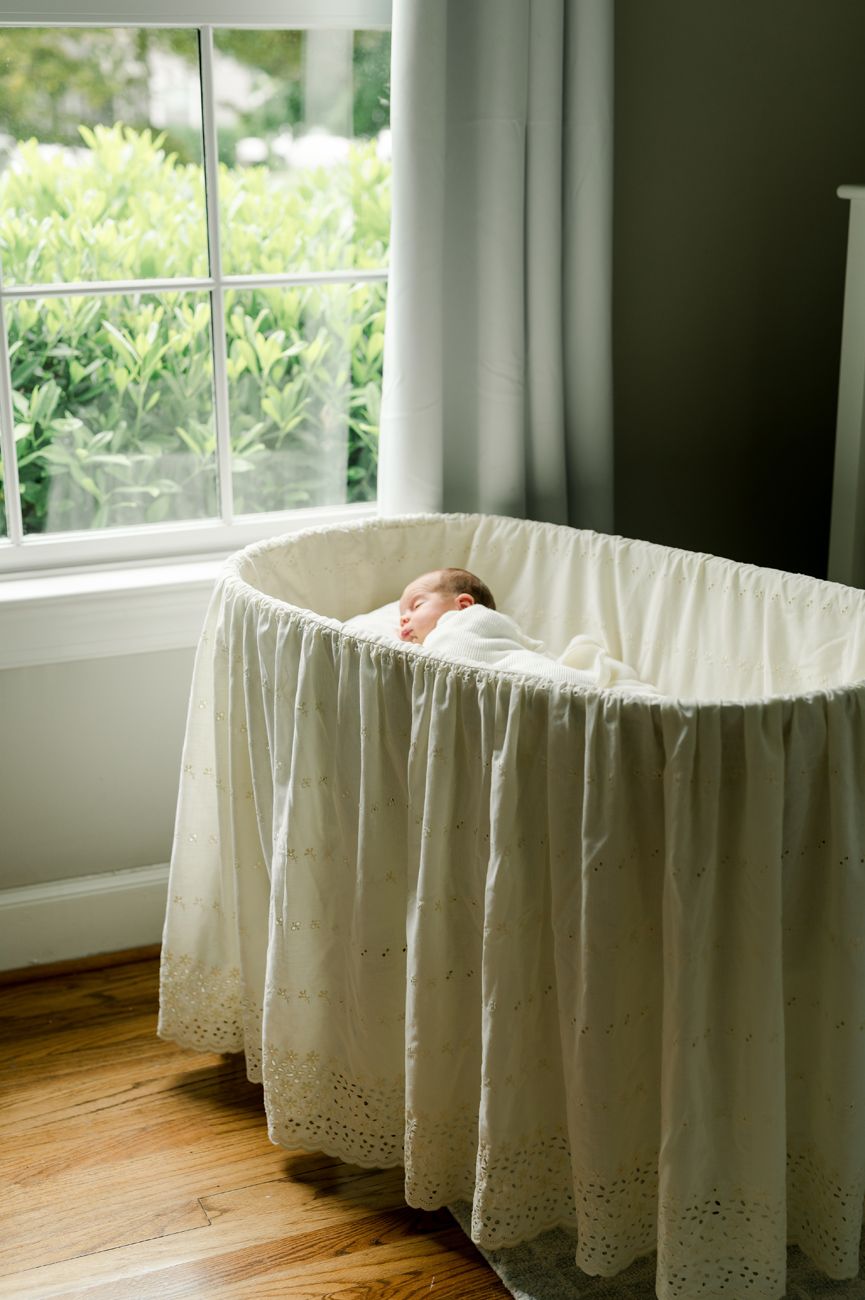 A white vintage bassinet with eyelet lace trim sits beside a sunlit window.