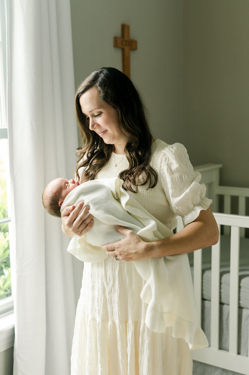A tender moment captured by a window as a parent in a white dress cradles a newborn baby with a cross on the wall.