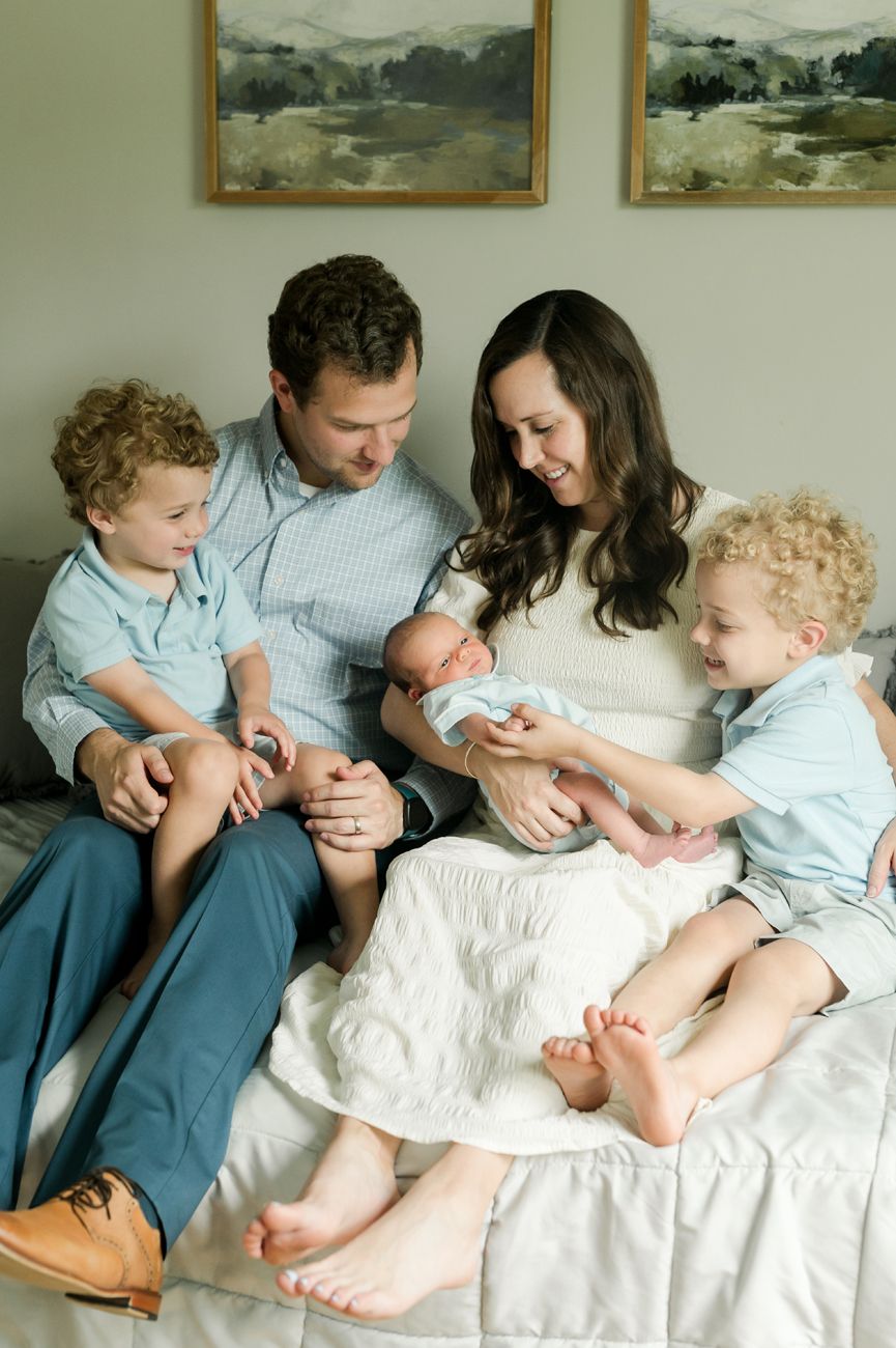 Family sits together on white bedding as they welcome newborn baby with two toddler siblings.