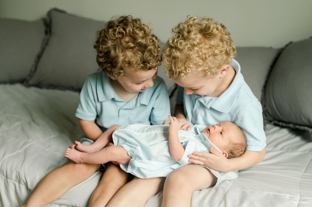Two young children in matching blue shirts sit on a gray couch holding their newborn sibling.