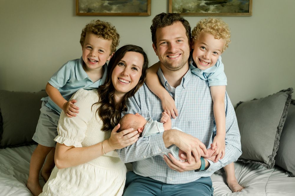 A loving family relaxes together on a grey couch, with twin boys in matching blue outfits and a newborn baby.