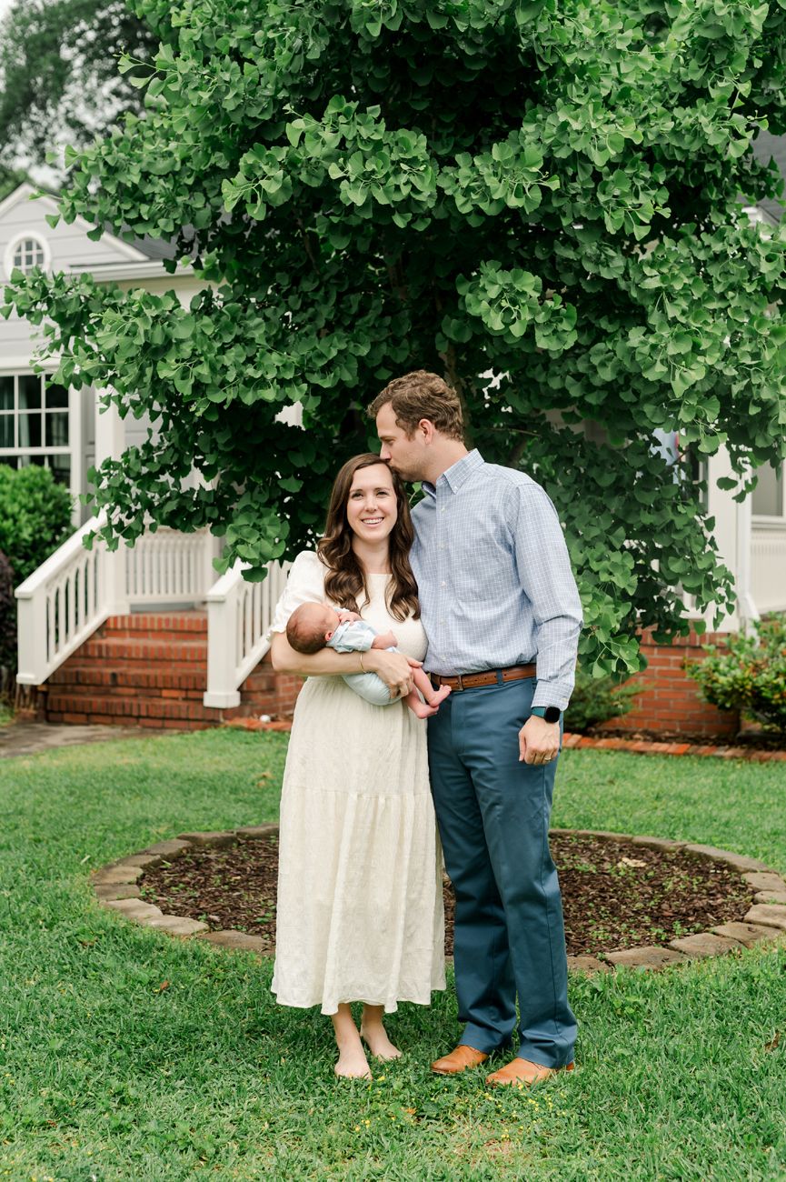 A couple stands together on a lawn holding a baby in front of a white house with brick steps and lush greenery.