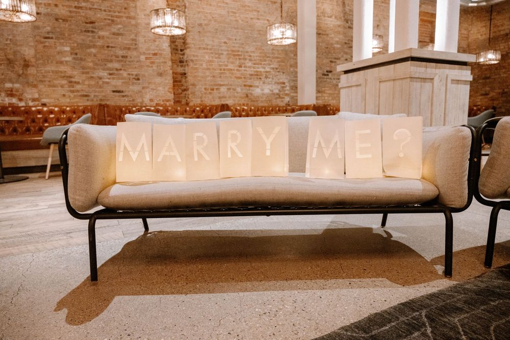 A vintage metal bench with 'MARRY ME?' written in rustic letters on its cushion inside a brick-walled space with chandeliers.