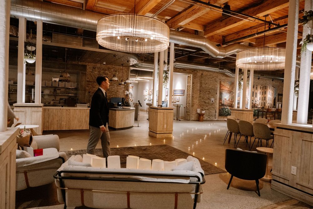 Elegant event space features exposed brick walls, circular chandeliers, and modern furnishings in a rustic-industrial setting.