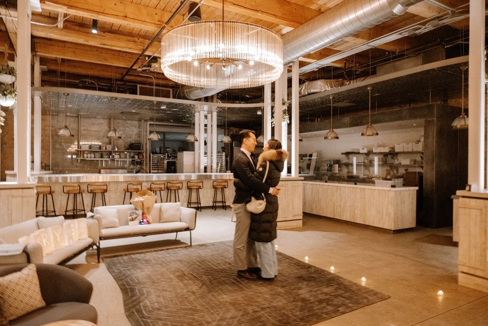 Elegant modern industrial event space with grand chandelier, exposed beams, and couple dancing in warm lighting.