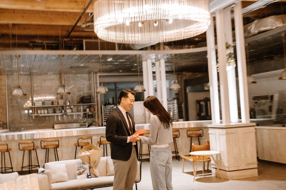 Two individuals in formal attire share a moment in an upscale restaurant interior with modern lighting and bar seating.