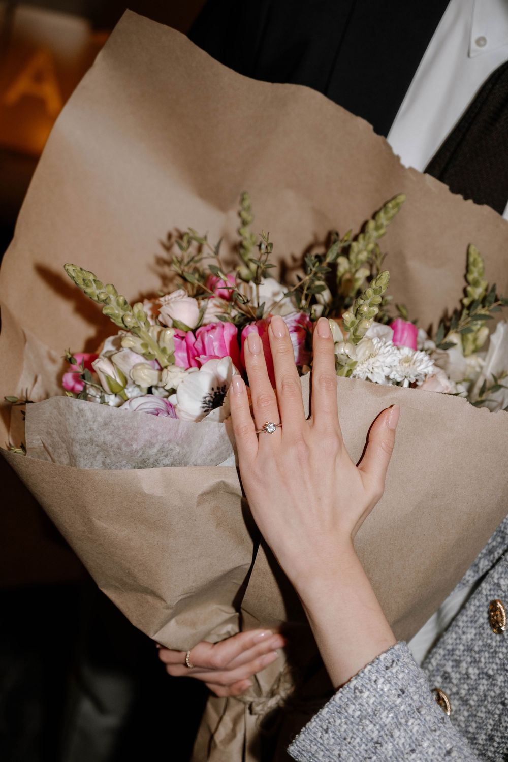 A hand wearing a gold ring holds a romantic bouquet wrapped in brown paper featuring pink roses and white flowers.